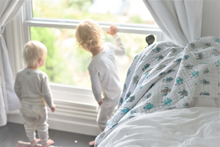Two toddlers looking out a window beside a Cotton baby blanket on the bed, featuring playful designs.