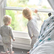 Two toddlers looking out a window beside a Cotton baby blanket on the bed, featuring playful designs.