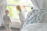 Two toddlers looking out a window beside a Cotton baby blanket on the bed, featuring playful designs.