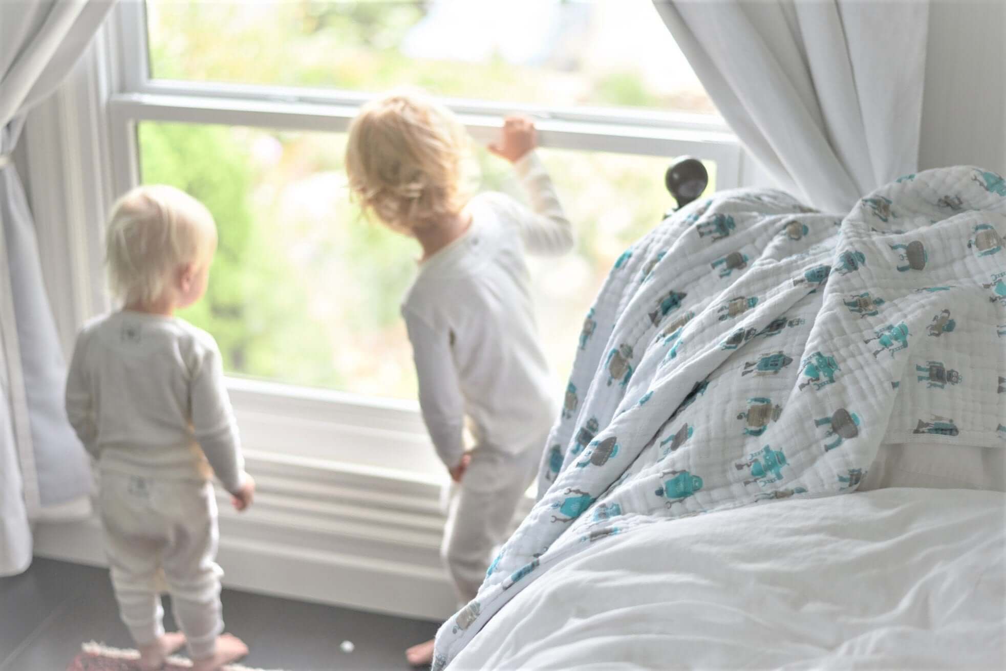 Two children looking out the window beside a cozy Cotton Baby Blanket draped over a bed.