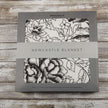 American Rose Newcastle Blanket packaging with floral design and brand logo on a rustic wooden background.